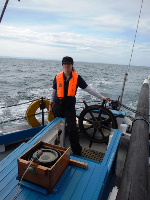 Catherine at helm of Reaper. She is wearing a Breton hat, a navy polo shirt and an orange life jacket. She is holding onto the wheel of the boat.