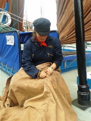 Catherine is sat repairing a large brown sail. She is wearing a navy boiler suit embroidered with 'Catherine Reaper FR358', a red neck scarf and a navy Breton.