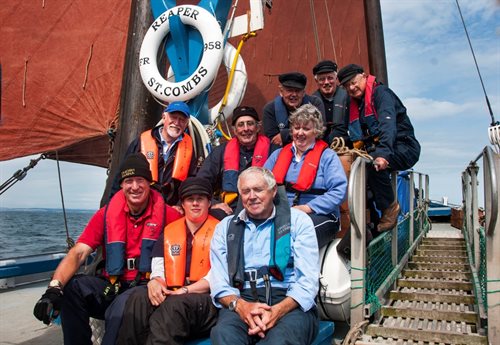 Nine members of the Reaper crew. They are sat on the Reaper in front of a brown sail. Catherine is sat in the middle front row.