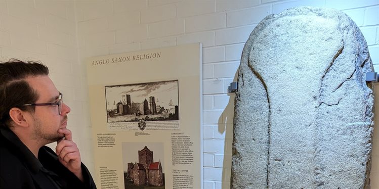 Curating Visibility Fellow Karl Mercer - a white male in his mid-30s, dark brown hair in a side-parting, with a short beard and glasses wearing a dark shirt and jacket - is looking at a carved stone slab, a tomb marker - propped up against a white-washed brick wall.   An interpretation board is between them titled "Anglo Saxon Religion" showing images of the Anglo Saxon church of St Mary in Castro.